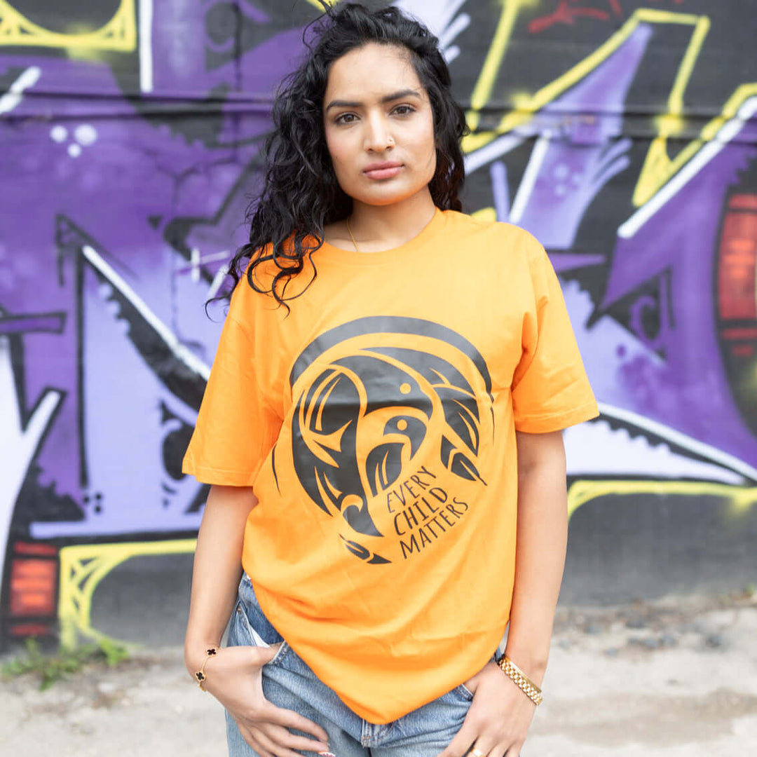 Woman wearing a every child matters t-shirt with a graphic design in front of a graffiti-covered wall.