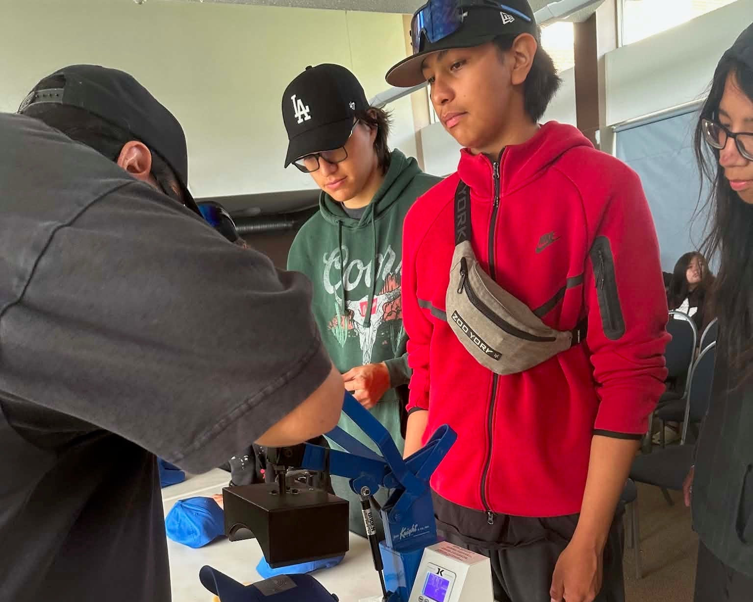 A group of people gathered around a blue and white heat press machine on a folding table in an indoor workshop or classroom setting.
