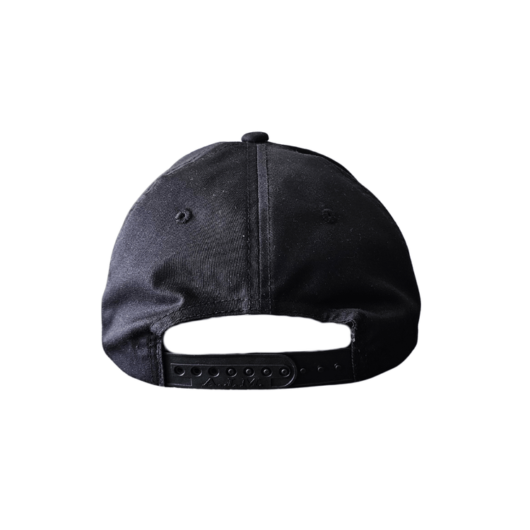 Black snapback hat with white printed Red Rebel Armour crest on front panel, displayed on a wooden table facing backwards.