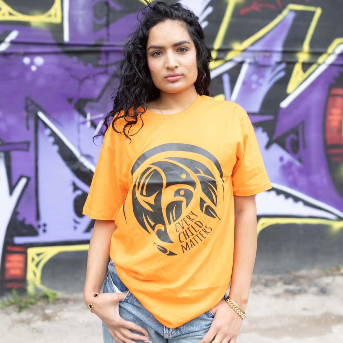 Woman wearing a every child matters t-shirt with a graphic design in front of a graffiti-covered wall.