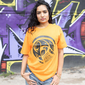 Woman wearing a every child matters t-shirt with a graphic design in front of a graffiti-covered wall.