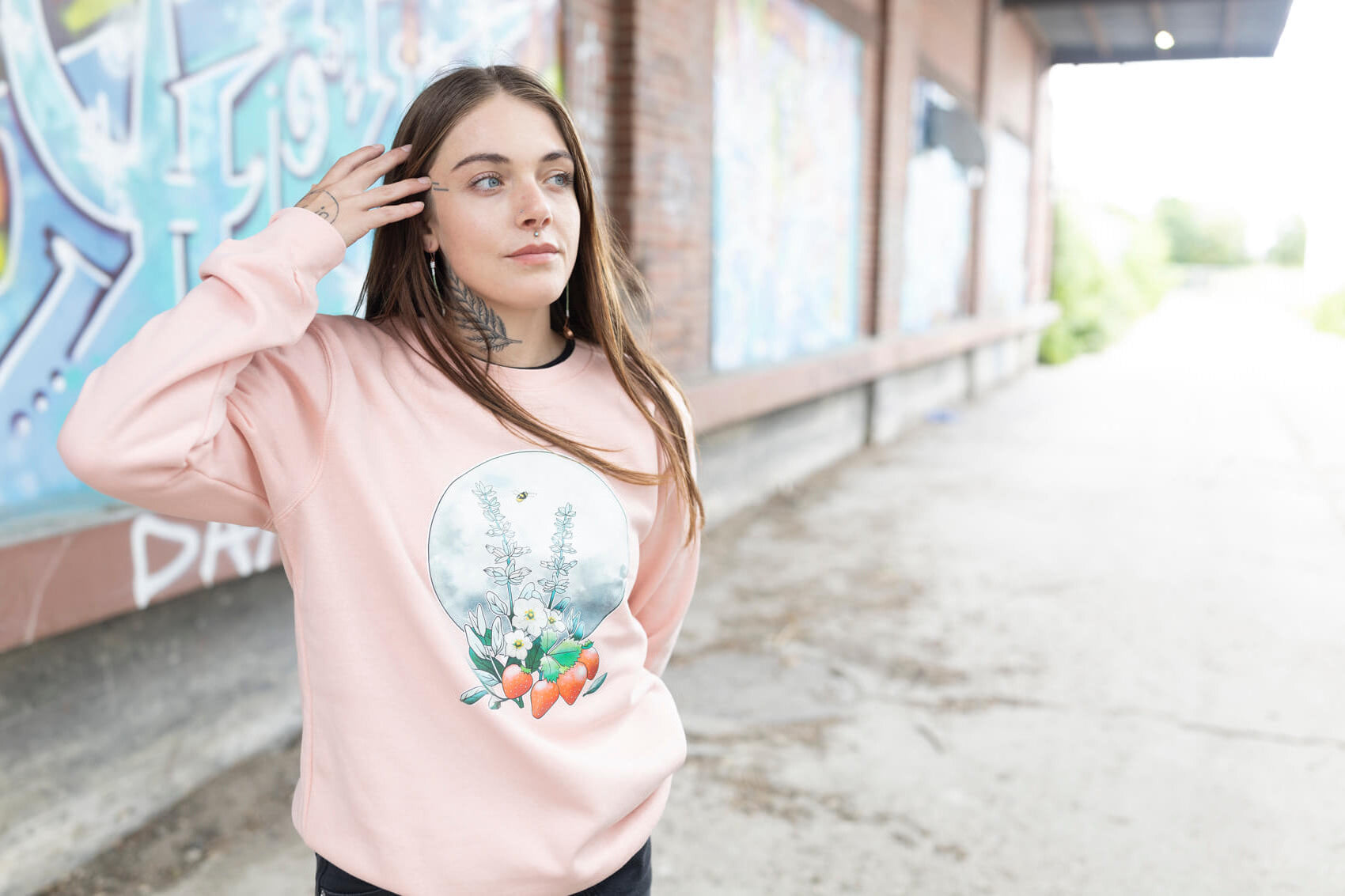 Person wearing a light pink sweatshirt with a graphic design, standing on a street with graffiti in the background.