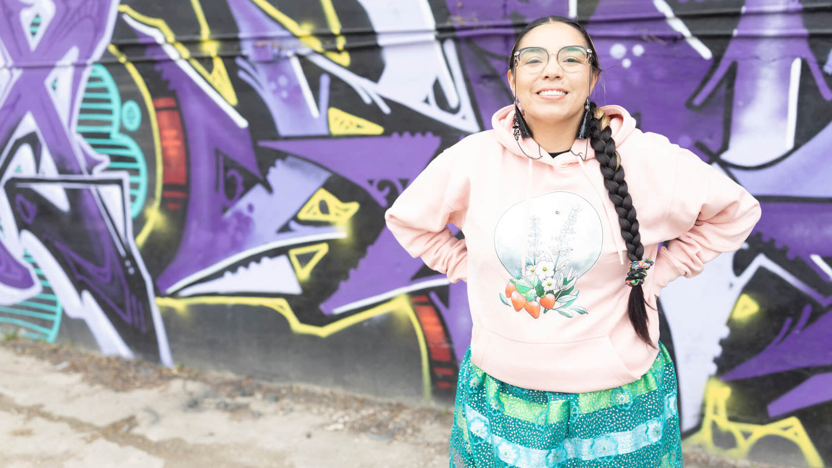 Person wearing a pink hoodie with a colorful design, standing in front of a graffiti-covered wall.