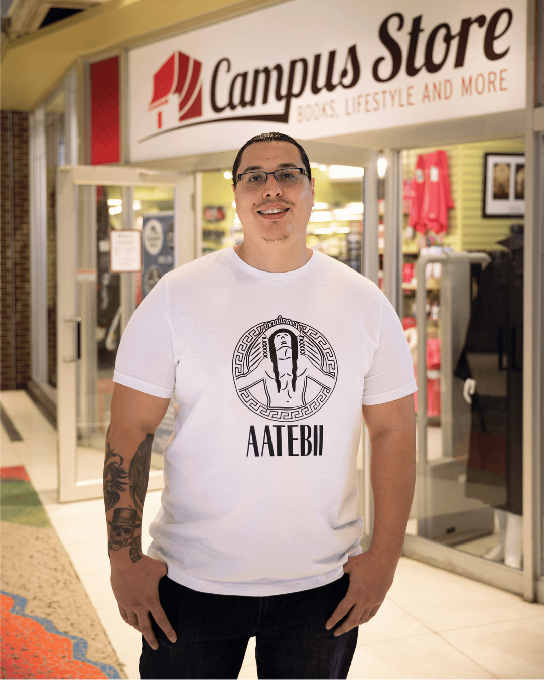A man wearing a white graphic t-shirt with a black circular design and the text 'AAATEBI' stands in front of a storefront sign that reads 'Campus Store'.